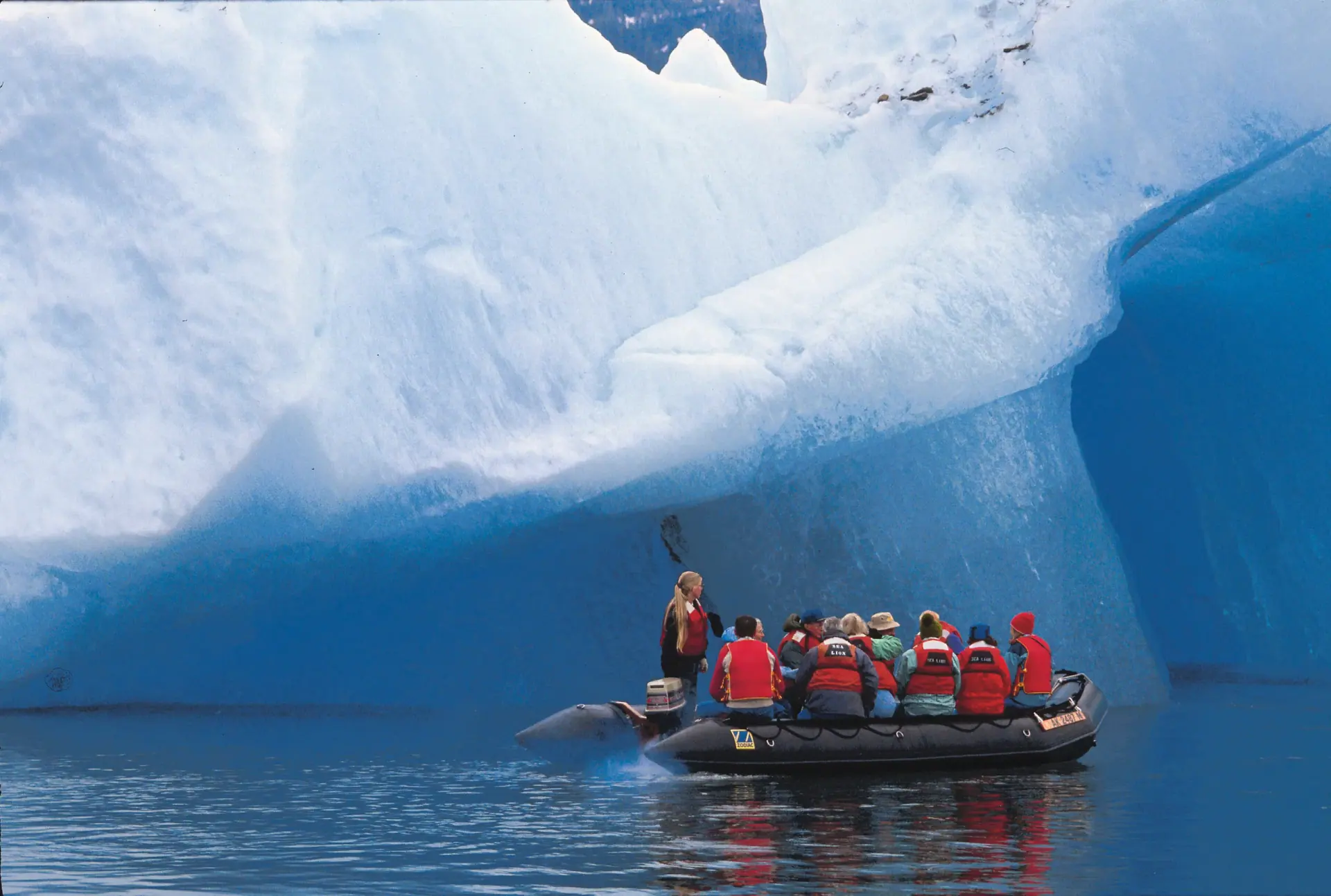 Expedition guests in a Zodiac exploring beneath a massive blue iceberg