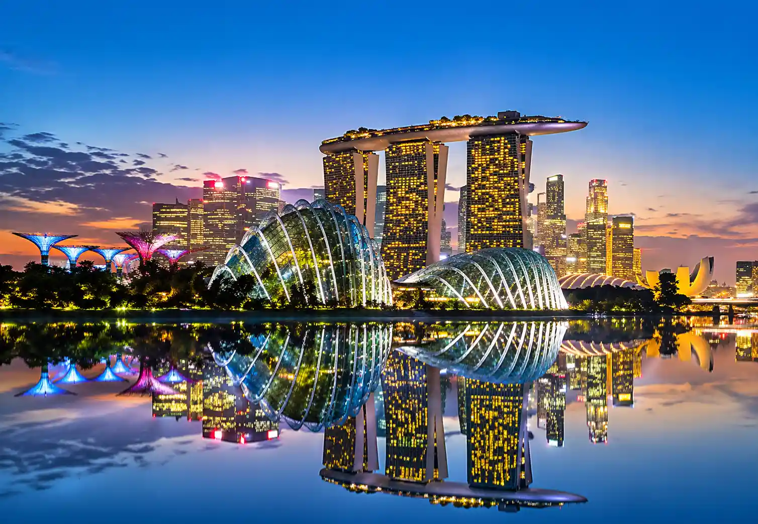 Singapore skyline at dusk featuring Marina Bay Sands, Gardens by the Bay, and illuminated waterfront reflections.